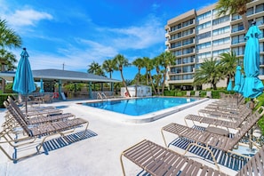 A pool area with lounge chairs and umbrellas, a building with balconies, and palm trees.