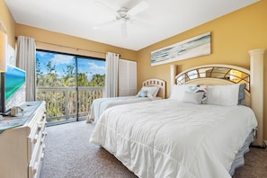 A hotel room with two beds, a ceiling fan, a large window with curtains, and a view of greenery outside.