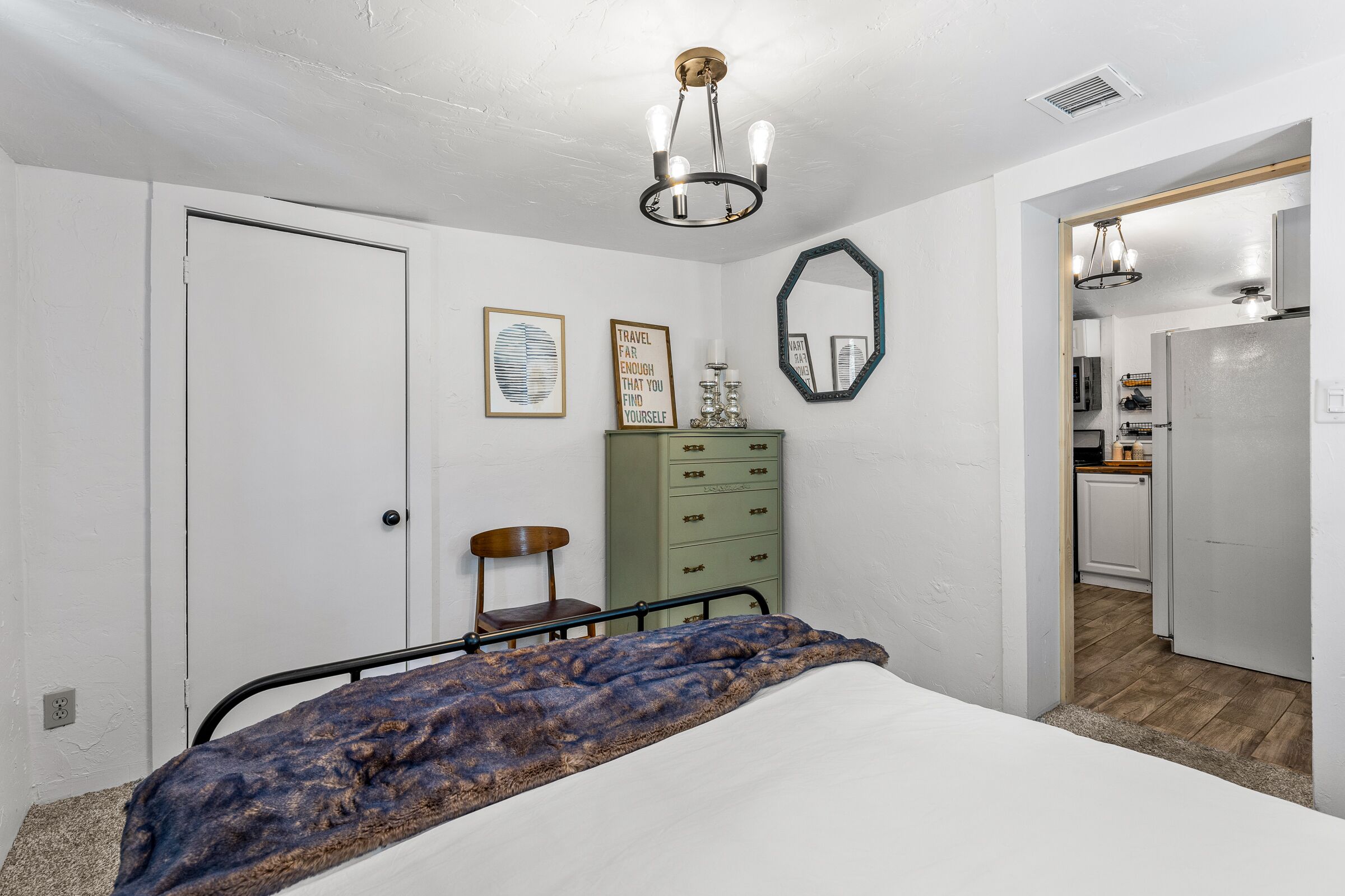 Un dormitorio con cama, cómoda, espejo y puerta que da a una cocina.