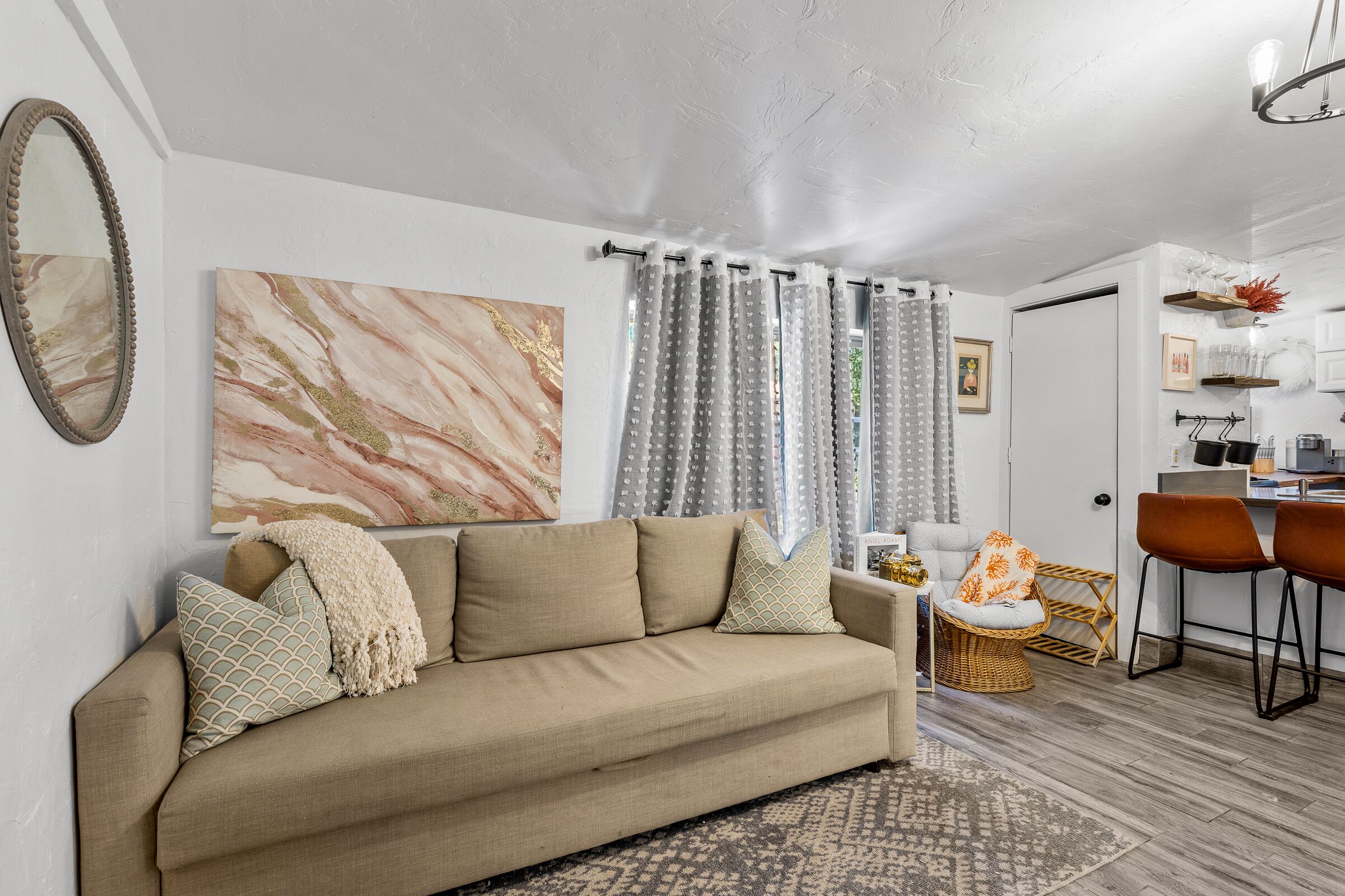 A living room with a beige sofa, patterned cushions, a large abstract painting, and a dining area with a wicker chair and a small round table.
