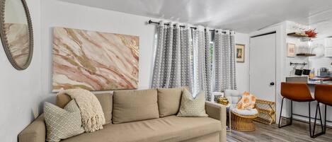 A living room with a beige sofa, patterned cushions, a large abstract painting, and a dining area with a wicker chair and a small round table.