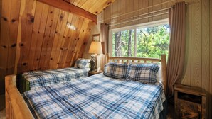 Dormitorio de cabaña de madera con dos camas, una mesita de noche y ventana con vista a árboles.