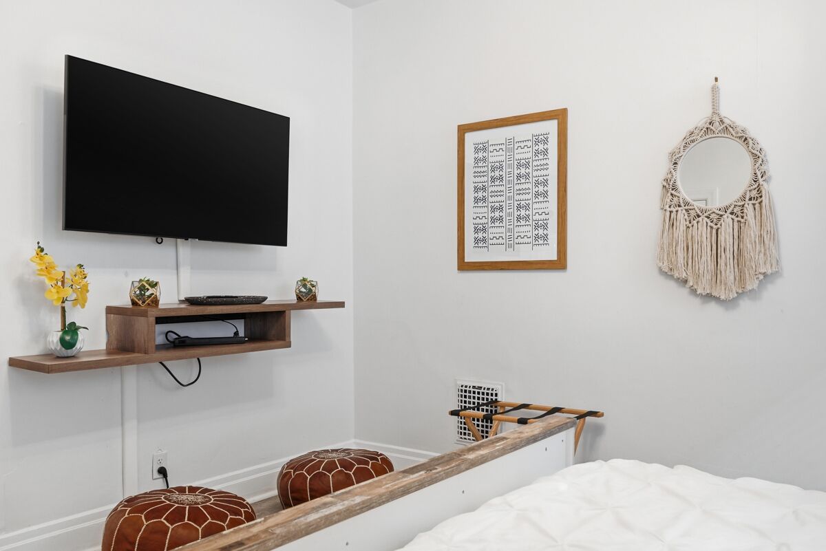 A bedroom with a bed, a TV mounted on the wall, a framed artwork, a macrame wall hanging, and a shelf with decorative items.