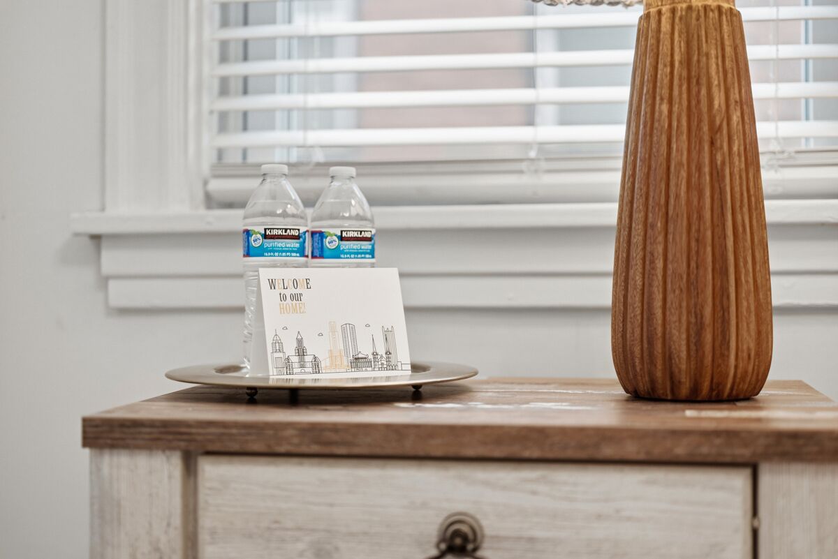 A wooden nightstand with a tray holding two bottles of Kirkland water, a welcome sign, and a tall vase.