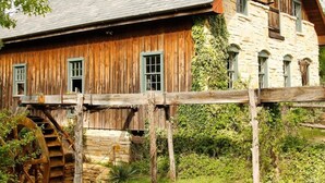 A rustic wooden building with a water wheel and stone walls, surrounded by greenery. - Oak Tree Manor w/ Pool (Waco)