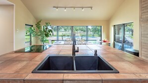 A kitchen with a large sink, a dining area with a table and chairs, and large windows offering a view of the outdoors. - Oak Tree Manor w/ Pool (Waco)