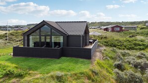 Una casa moderna de color oscuro con un amplio ventanal y un techo inclinado, rodeada de exuberante vegetación y otras viviendas al fondo.