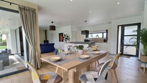 Un comedor moderno con una mesa de madera dispuesta para cuatro personas, rodeada de sillas estampadas. Se ve un sofá azul y una cocina con un amplio ventanal al fondo.