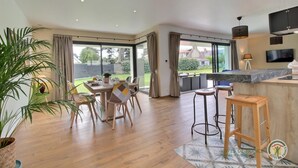 Un comedor amplio con una gran mesa y sillas, una zona de cocina con encimera de mármol y vistas al jardín a través de ventanas grandes.