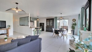 A modern living room with a grey sofa, a dining area with a round table and chairs, and a kitchen with dark cabinets and a black refrigerator. - Villa des Dîmes - Pleneuf VAL Andre (PLENEUF VAL ANDRE)