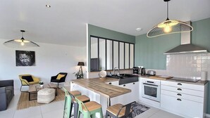 A modern kitchen with a central island, white cabinetry, and a built-in oven. There are bar stools and a dining table with chairs. - Villa des Dîmes - Pleneuf VAL Andre (PLENEUF VAL ANDRE)