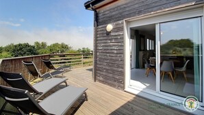 A wooden deck with lounge chairs and a glass door leading to an indoor dining area. - Villa des Dîmes - Pleneuf VAL Andre (PLENEUF VAL ANDRE)