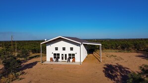 A white single-story building with a large front porch, a covered outdoor area, and a clear view of the surrounding landscape. - Gorgeous, Secluded Hill Country Escape Near River + Game room! (Llano)