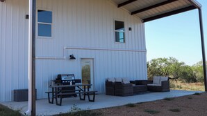 A white building with a covered patio area featuring a grill, picnic table, and wicker furniture. - Gorgeous, Secluded Hill Country Escape Near River + Game room! (Llano)