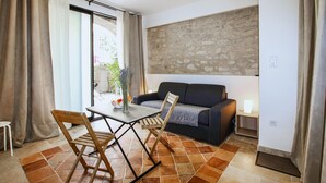 Uma sala de estar com sofá, mesa e uma cadeira. Apresenta uma parede de pedra e uma janela com cortinas.