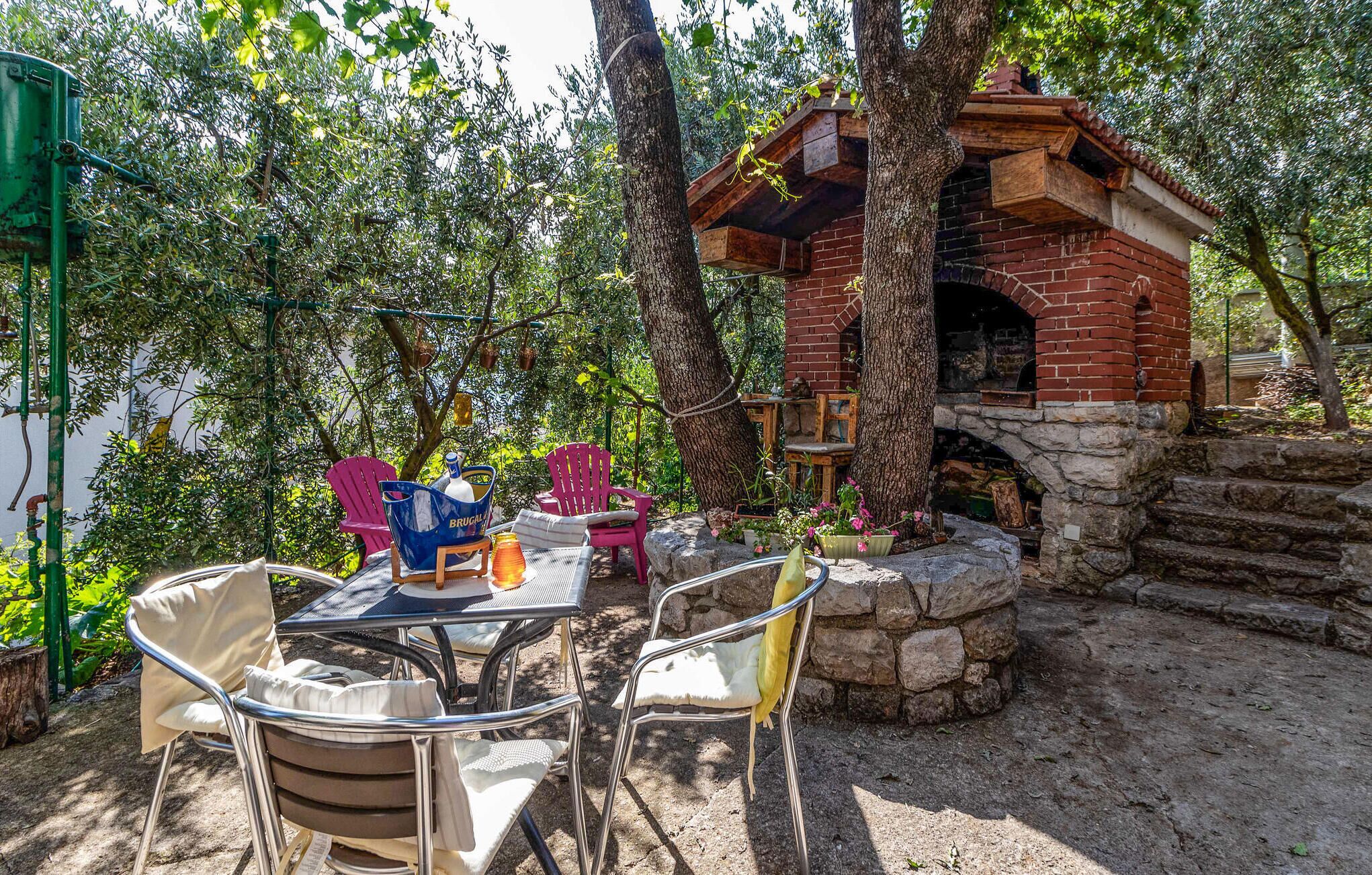 Un espace de restauration en plein air avec des chaises et une table en métal, entouré d’arbres et d’une structure en briques qui abrite un four.