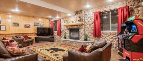 A cozy living room with leather sofas, a flat-screen TV, a stone fireplace, and a red arcade machine.