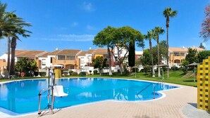 A swimming pool with a clear blue water, surrounded by a paved walkway. There are palm trees and a building with a tiled roof in the background.