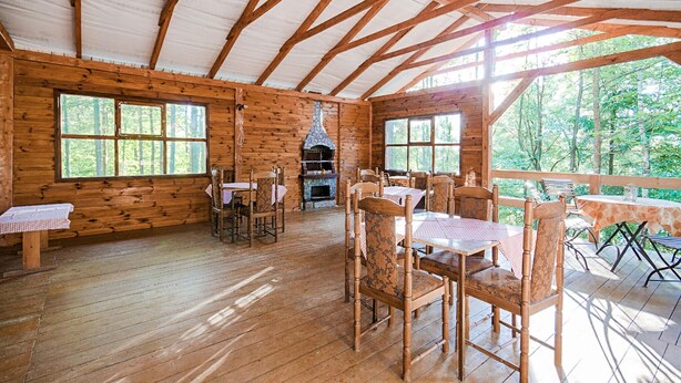 Un rustico chalet con pareti in legno e tetto di paglia. Dispone di un'area pranzo con sedie e tavoli in legno, un camino e ampie finestre che offrono vista sulla foresta circostante.