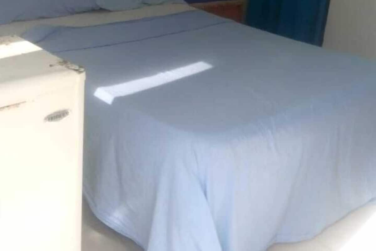 A single-bed room with a blue bedspread, a white wall, and a small table with a blue object on it.