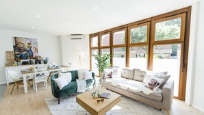 A modern living room with a green sofa, a beige sofa, and a wooden coffee table. There is a dining area with a white table and chairs, and a large window with wooden frames. - APPARTIO - Modern country-style vacation apartment for 6 people (Ölbronn-Dürrn)
