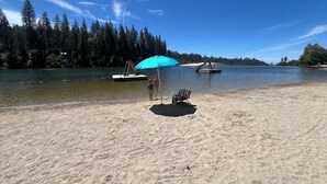 Una platja sorrenca amb una ombrel·la blava i una cadira, envoltada d'una zona boscosa i un llac tranquil.