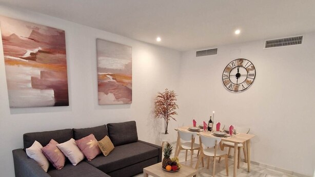 Una sala moderna con un sofá gris oscuro, arte abstracto en la pared, una mesa de comedor de madera con sillas y un gran reloj de pared.