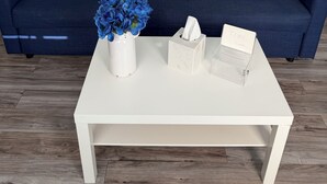 Una habitación con una mesa de centro blanca, un sofá azul, un florero con flores azules, una caja de pañuelos y un cuadro enmarcado de una isla tropical.