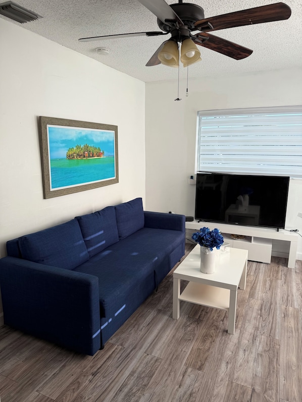 A living room with a dark blue sofa, a white coffee table with a vase of flowers, a ceiling fan, a large flat-screen TV, and a framed picture of a tropical island on the wall.