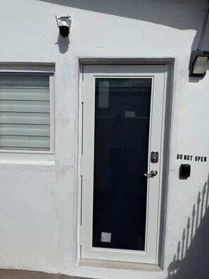 Eine weiße Tür mit Glasfenster, einem 'DO NOT OPEN' Schild, einer Überwachungskamera und einem Fenster mit Rollläden.