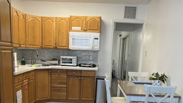 Cocina compacta con gabinetes de madera, microondas, tostadora y un lavabo. Hay un comedor con una mesa y sillas blancas, y un sofá azul.