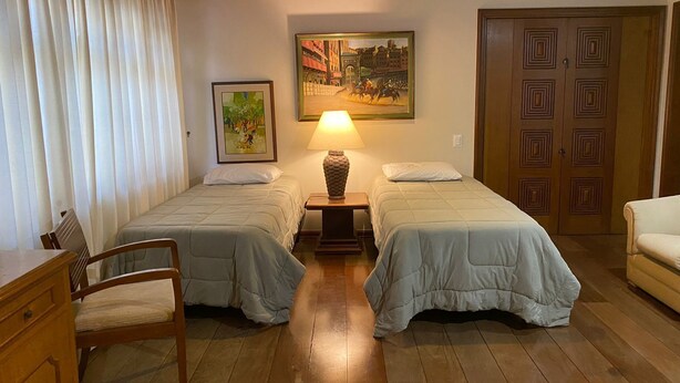 Une chambre avec deux lits simples, une commode en bois, une chaise, une lampe et une porte ornée d’un motif géométrique.