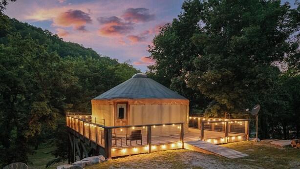 A wooden yurt with a fenced deck, surrounded by trees and a gravel path.