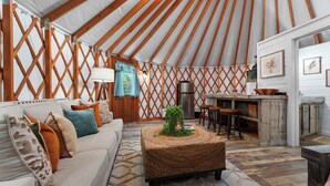 A cozy interior of a yurt with a sofa, a woven ottoman, a kitchen area, and a dining table.