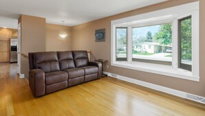 Een woonkamer met een bruine leren bank, een eettafel en een raam met uitzicht op een woonwijk.