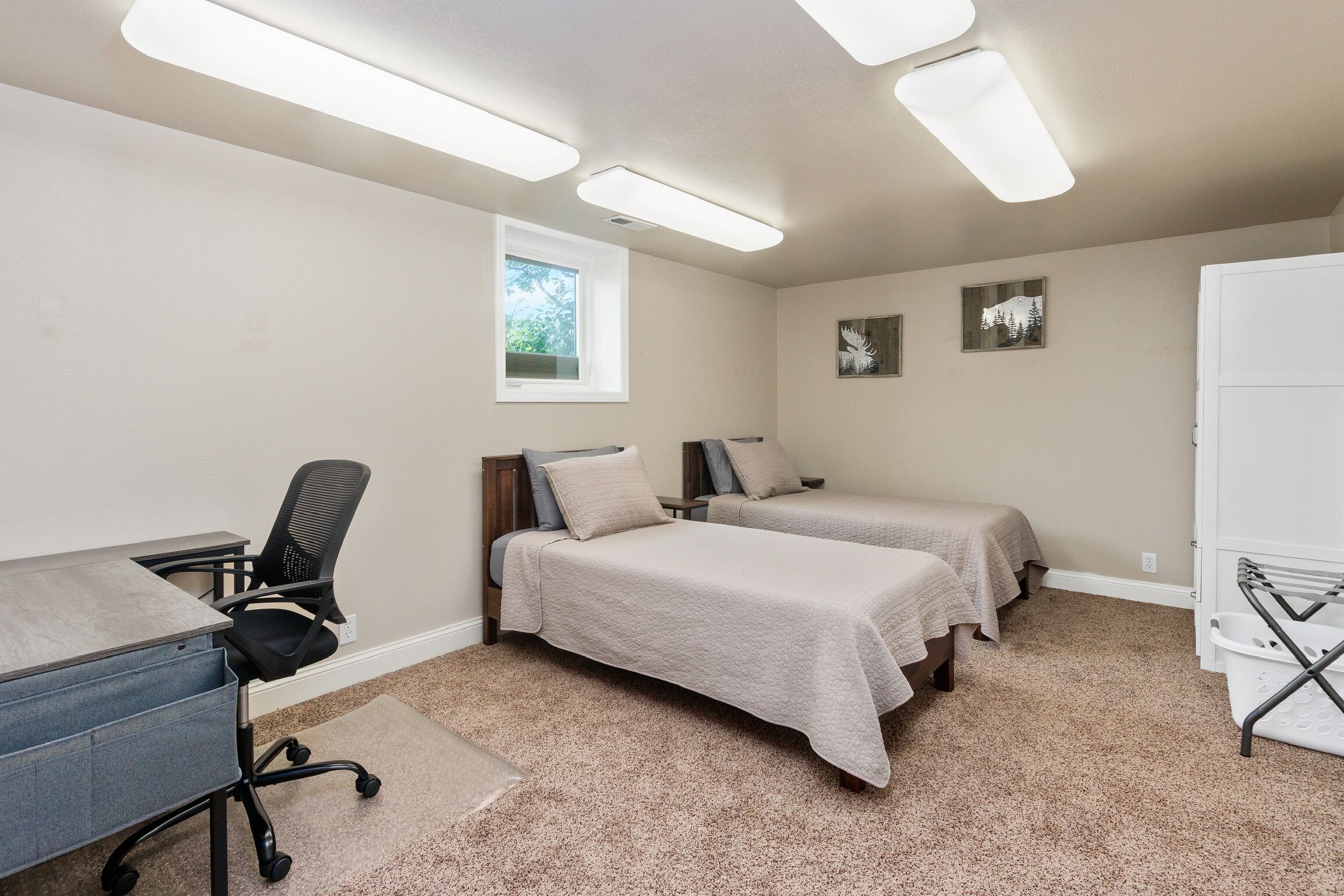 A room with two beds, a desk with a chair, a window, and a wall-mounted shelf.