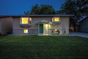 A two-story house with a green door, two windows with illuminated interiors, and a small front yard with trimmed grass and two potted plants. - Centrally located, huge shaded patio with fire pit! Entire place to yourself! (Billings)