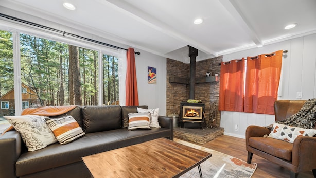 A modern living room with a fireplace, leather sofa, and a large window overlooking a forest. - Vanderhut Chalet (Sugarloaf)