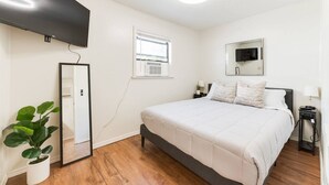 Un dormitorio con cama, televisor, ventana, espejo, planta y ventilador de techo.