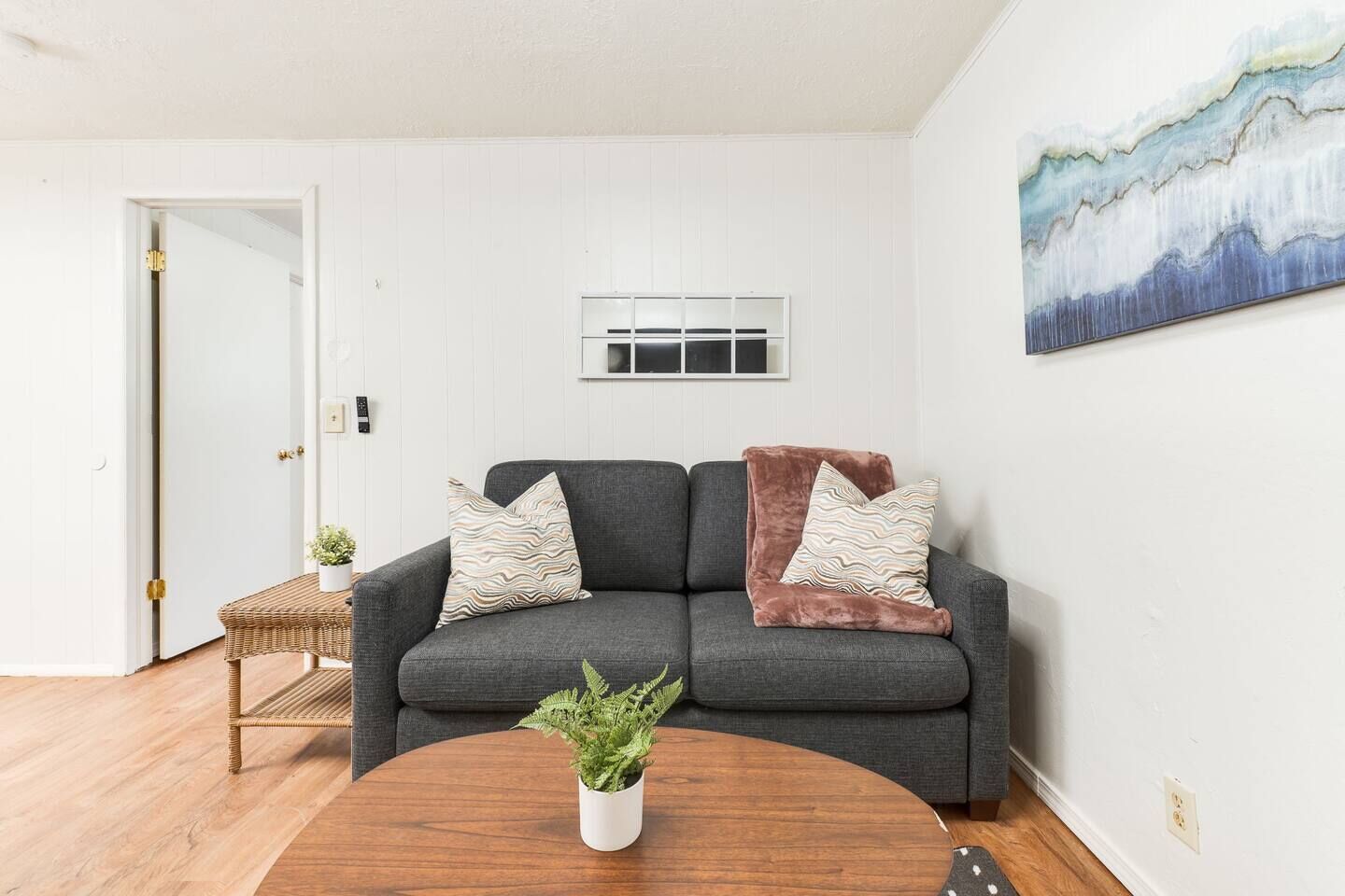 A living room with a grey sofa, two patterned cushions, a small side table with a plant, and a painting on the wall.