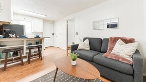 Una sala de estar moderna con un sofá gris, una mesa de centro de madera y una zona de cocina con armarios blancos y una mesa de comedor.