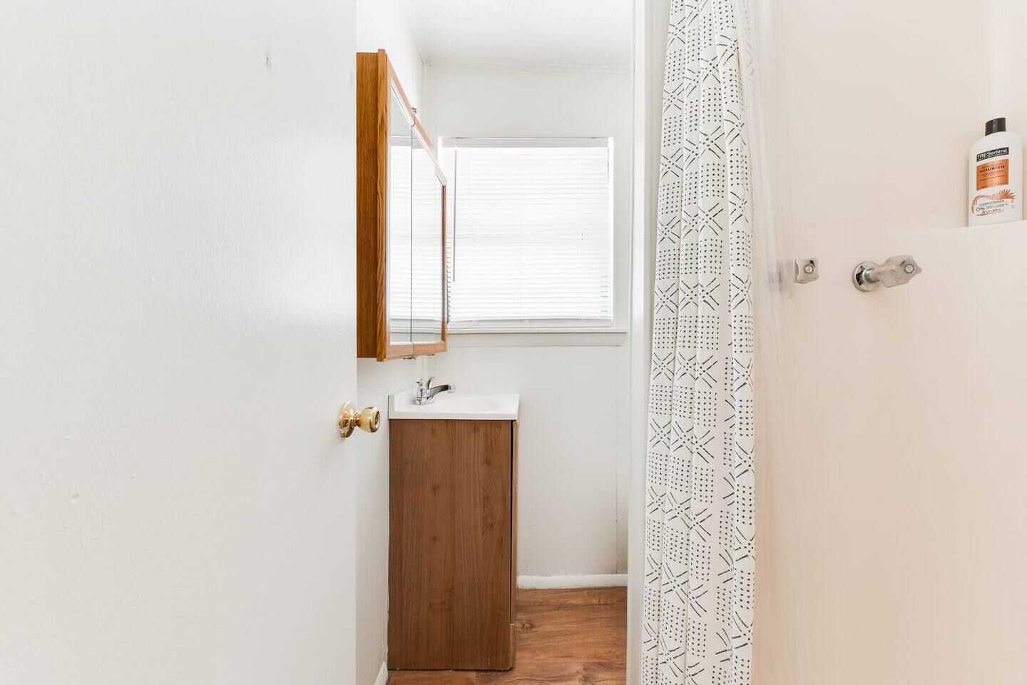 Ein schmaler Flur, der zu einem Badezimmer mit Holzschrank, Spiegel und einem Fenster mit Jalousien führt.