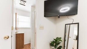 Una habitación con televisión montada, un espejo, una planta en maceta, un armario de madera y una ventana con persianas.