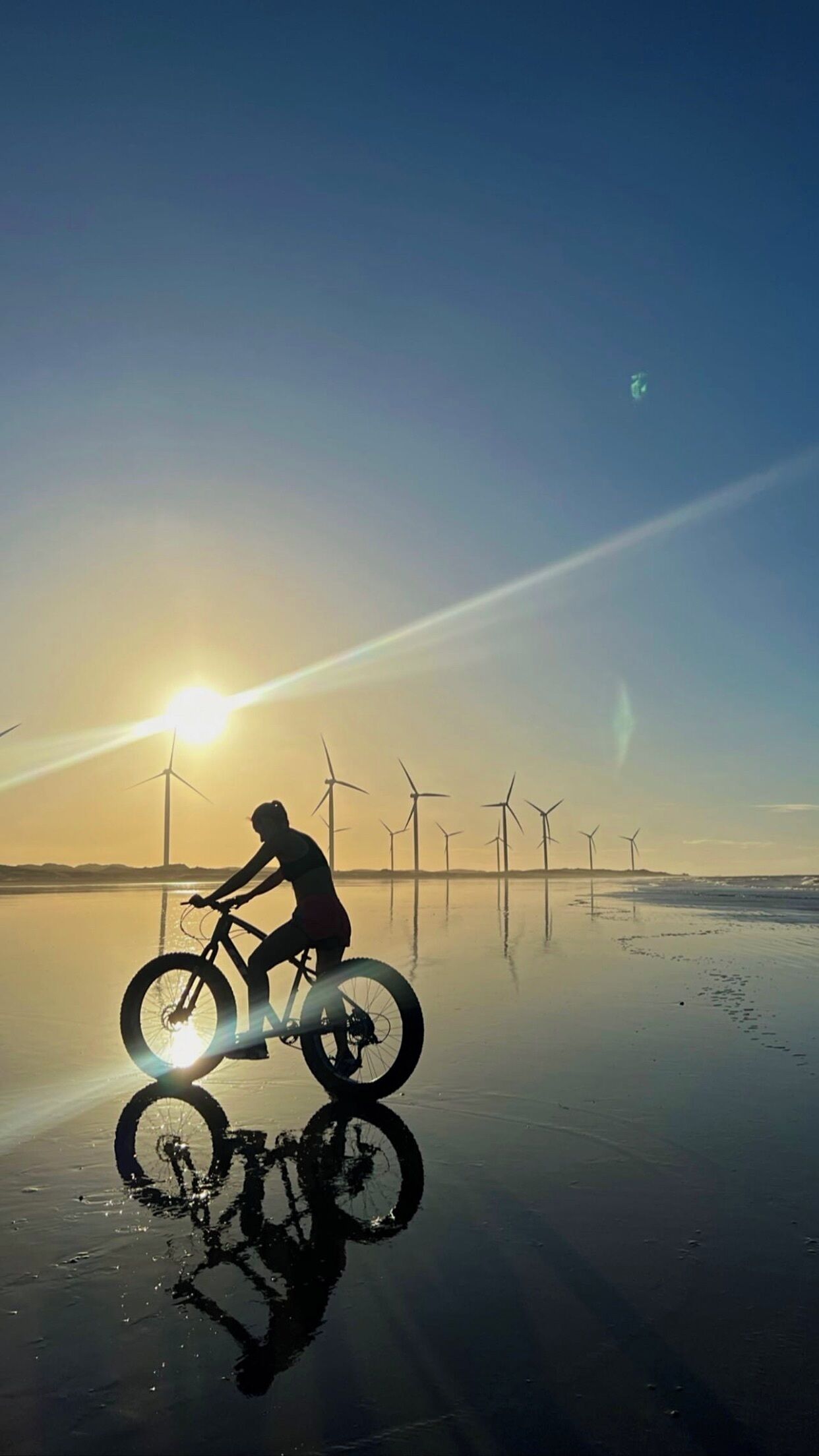A person riding a bicycle on a reflective water surface with wind turbines in the background during sunset.