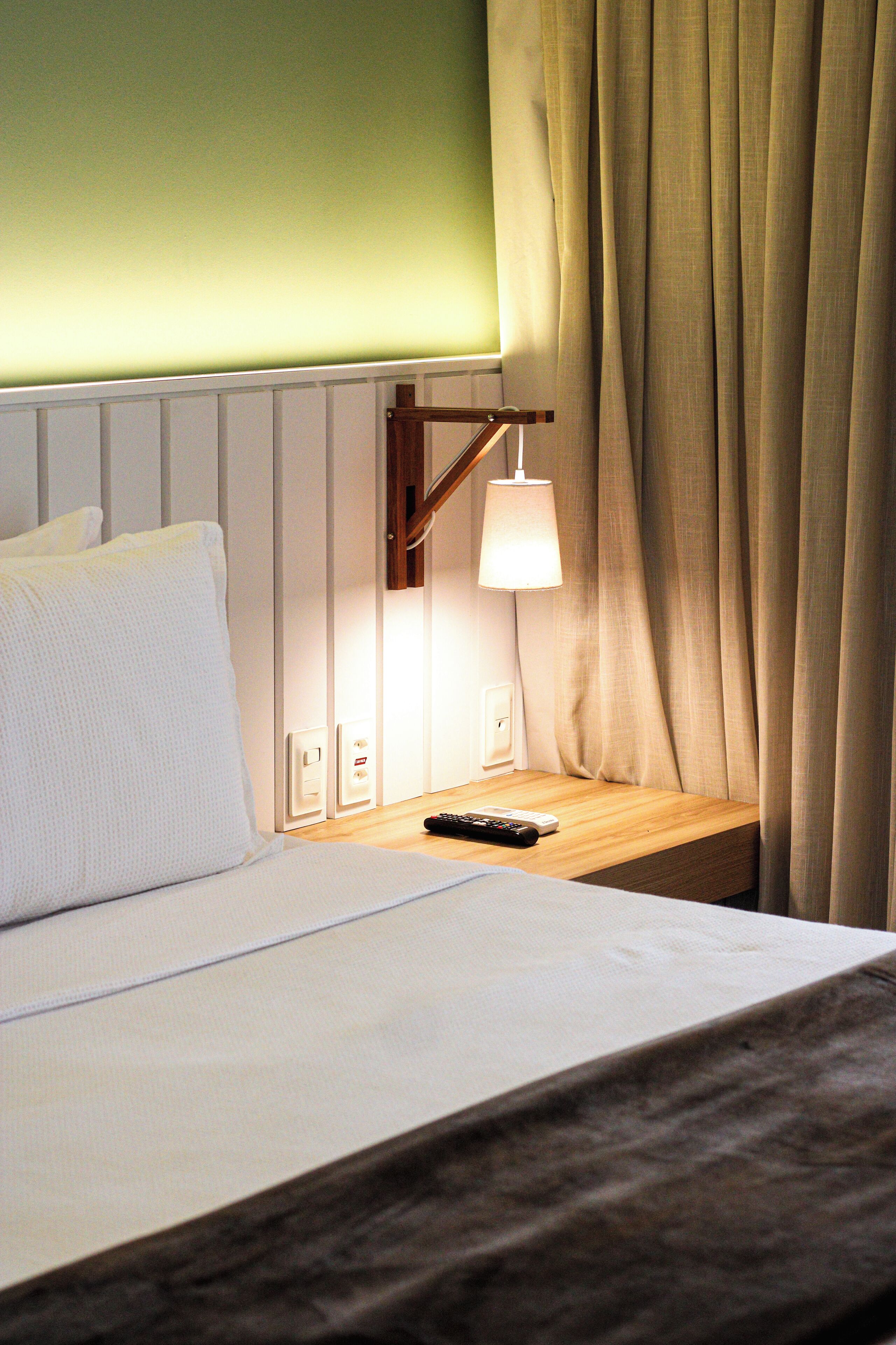 A neatly made bed with white linens, a bedside lamp, and a remote control on a wooden nightstand.