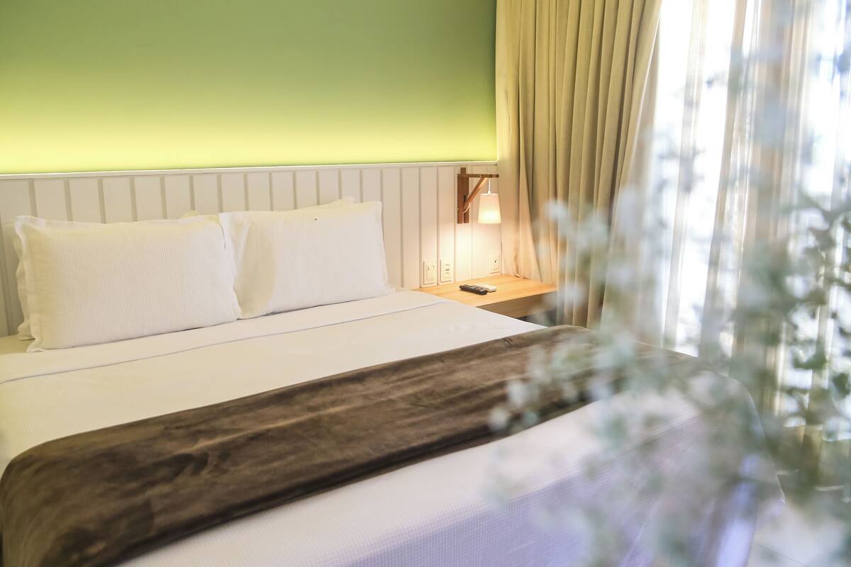 A hotel room with a neatly made bed, a bedside table with a lamp, and a view of the outdoors through the window.