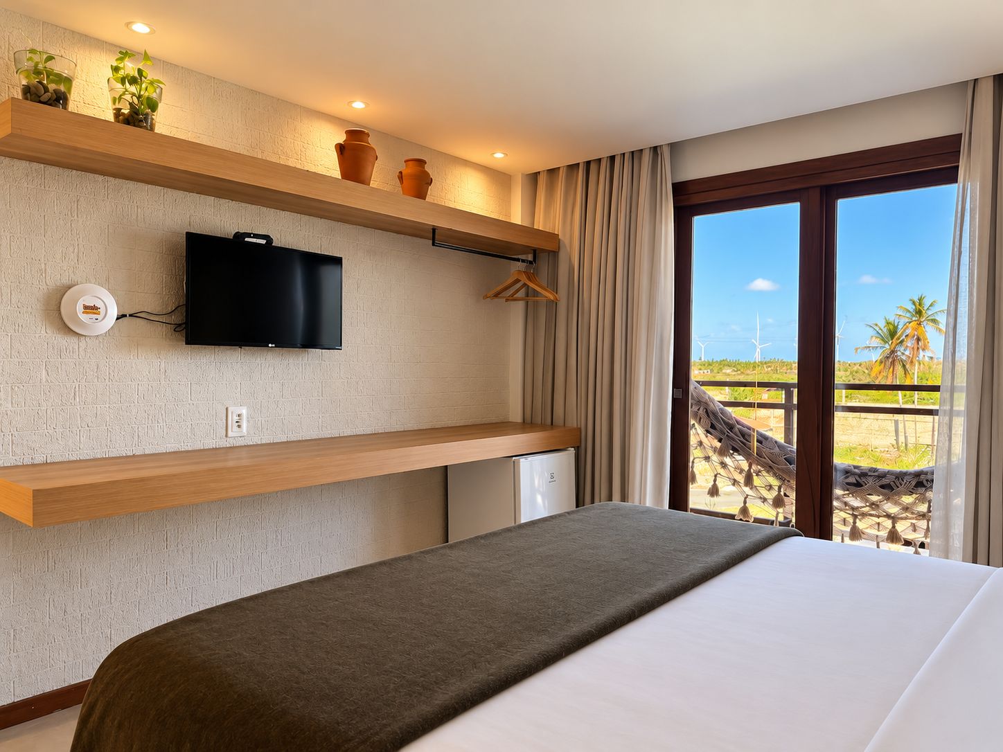 Une chambre d’hôtel avec un lit, une télévision, un petit réfrigérateur, une étagère avec des objets décoratifs et une vue sur la plage.