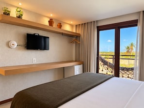 A hotel room with a bed, a TV, a small fridge, a shelf with decorative items, and a view of the beach. - HOTEL ICARAI BEACH (Amontada)