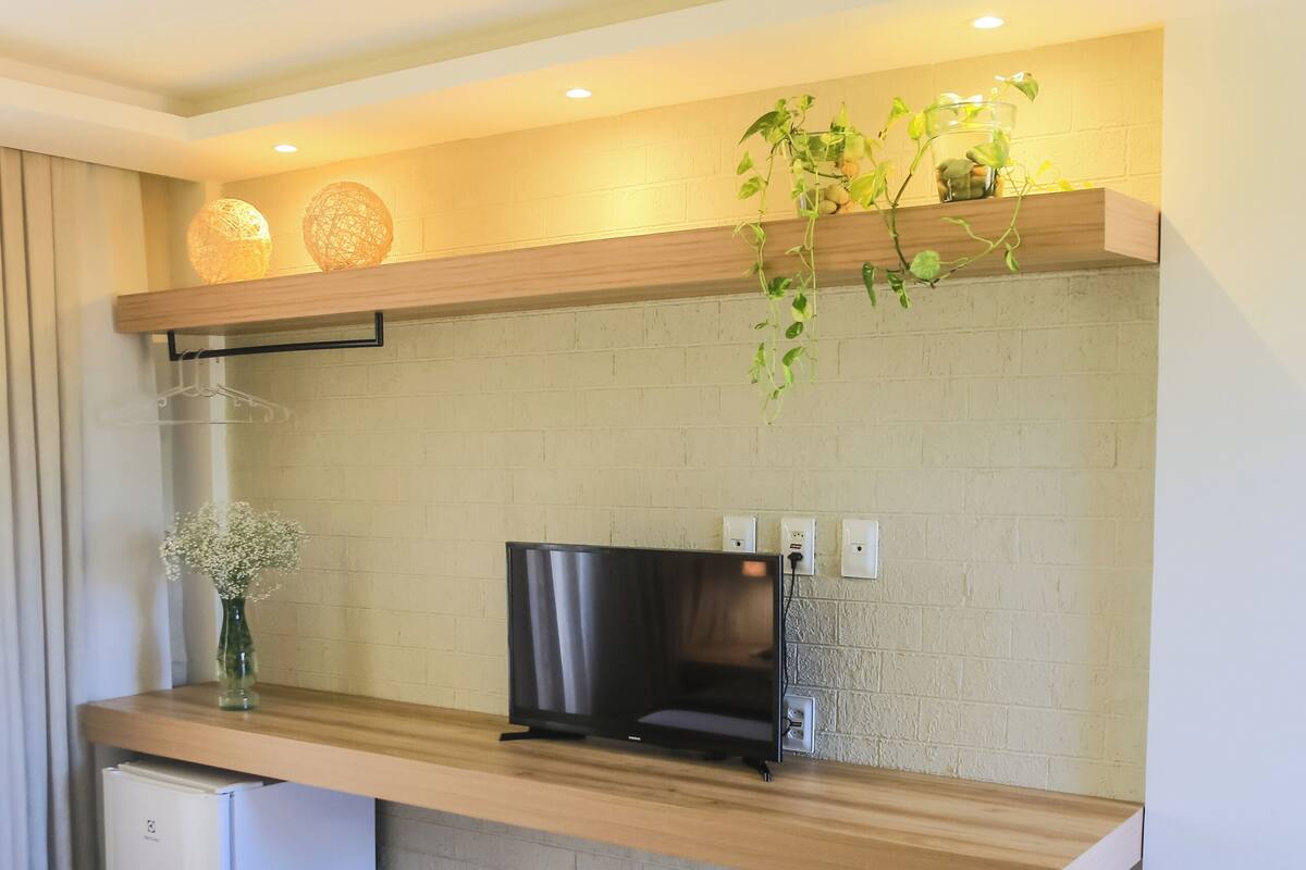 A modern hotel room with a wooden console table, a flat-screen TV, a small refrigerator, and a potted plant.