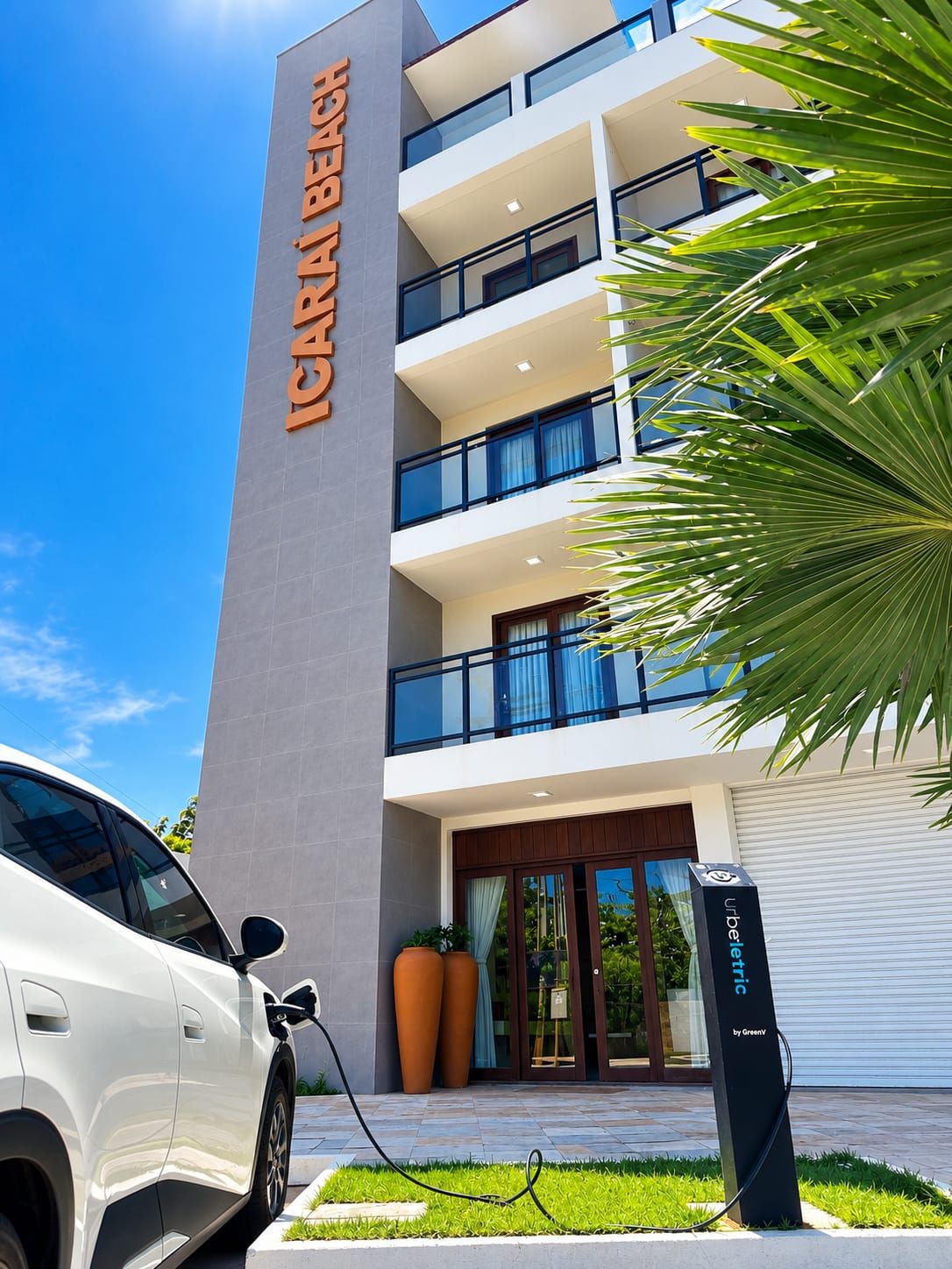 Een modern hotelgebouw met een opvallend bord, een laadpaal voor auto's en een palmboom.